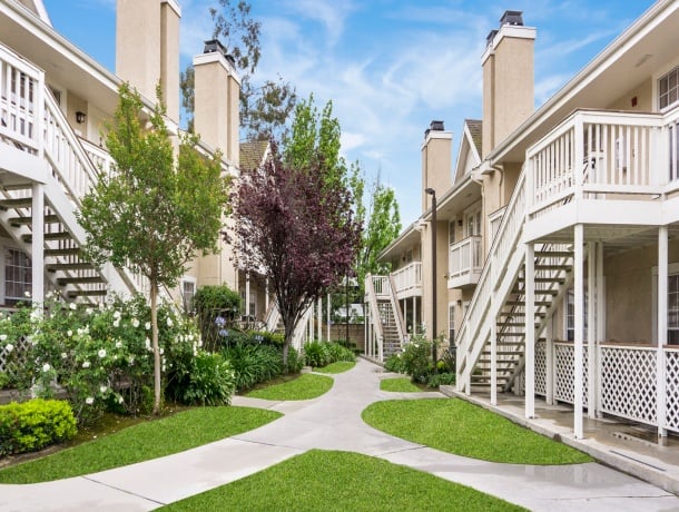 A courtyard with a beige building, white balconies, and staircases, surrounded by trees and grass.