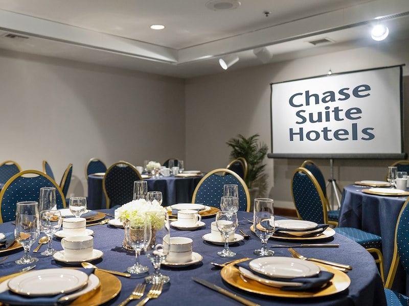 A formal dining setup in a meeting room with a projector screen displaying 'Chase Suite Hotels'.